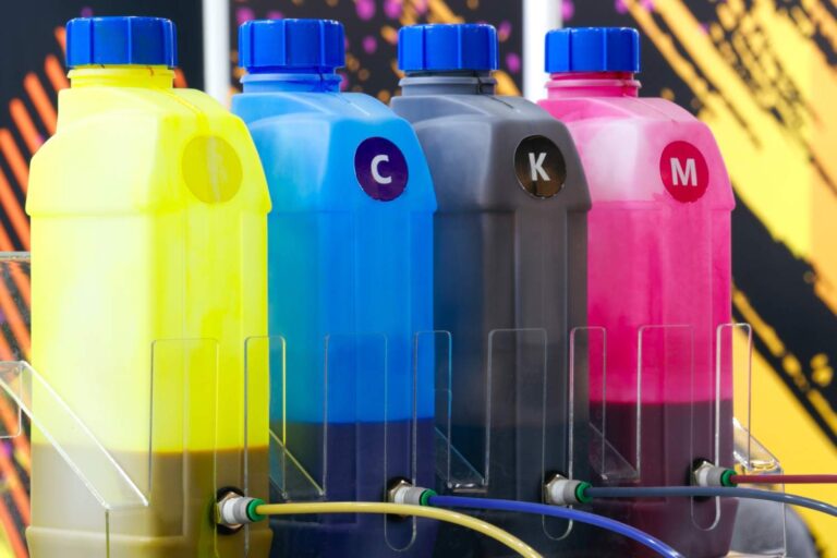 Four printer ink containers in yellow, cyan, black, and magenta (CMYK colors) with blue caps, arranged in a row on a clear holder with tubes attached